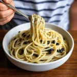 A close-up of a bowl of squid ink pasta with black squid ink pearls, served in a white bowl and twirled around a fork.