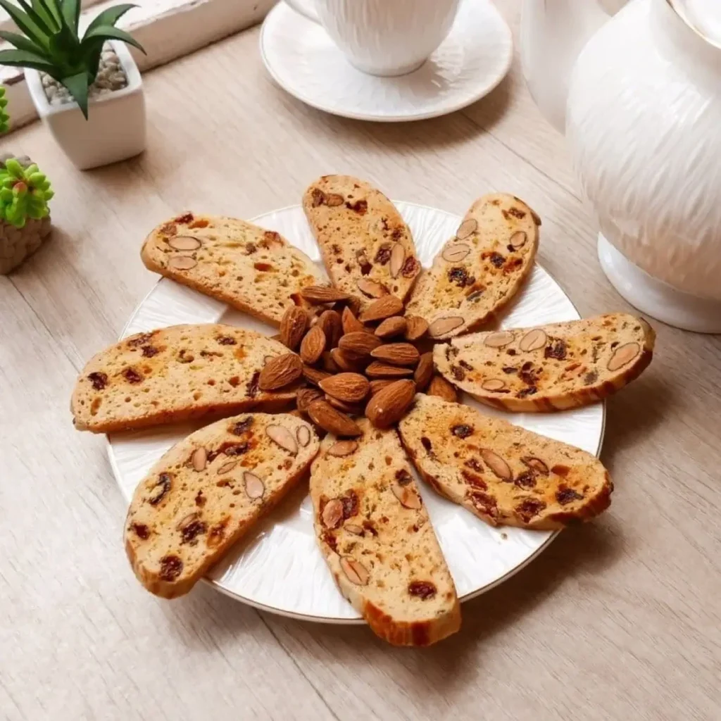 A plate of Moroccan Fekkas slices with almonds and raisins arranged in a floral pattern, with whole almonds in the center, set on a light wooden table with white tea ware.