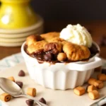 A warm and gooey chocolate cobbler topped with golden biscuits and a scoop of vanilla ice cream, served in a white ramekin. Chocolate chips and caramel pieces are scattered around.